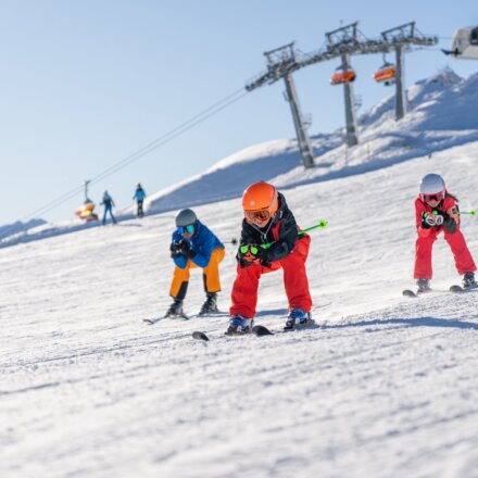 Pistenskifahren in Flachau. Am Bild sieht man: eine Familie die in der Hocke hinunterfährt. Im Hintergrund erkennt man die Skistation Starjet 3.