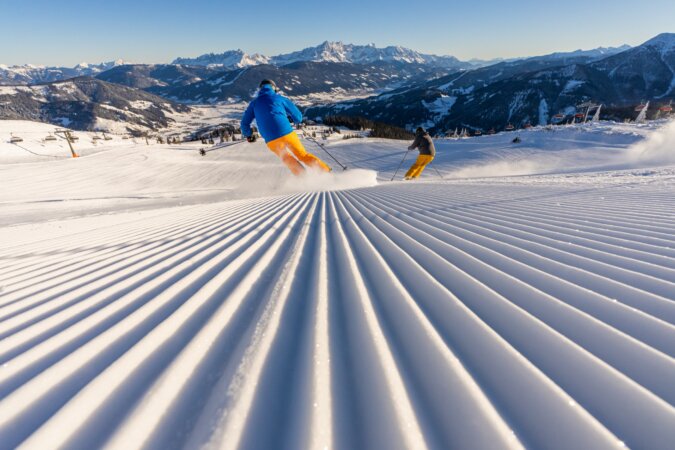 Pistenskifahren in Flachau. Am Bild sieht man: 2 Skifahrer von hinten auf der weiten frisch präparierte Piste mit Rillen, der herrliche Ausblick auf das Dachsteinmassiv, die Lifte Starjet 3 und Space Jet 3