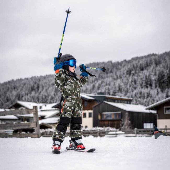 Kleines Kind mit Skihelm fährt Ski und hält Skistöcke im Schnee hoch