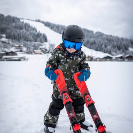 Kleines Kind mit Skihelm und Skibrille hält Kinderski im Schnee