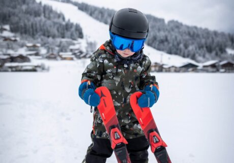 Kleines Kind mit Skihelm und Skibrille hält Kinderski im Schnee