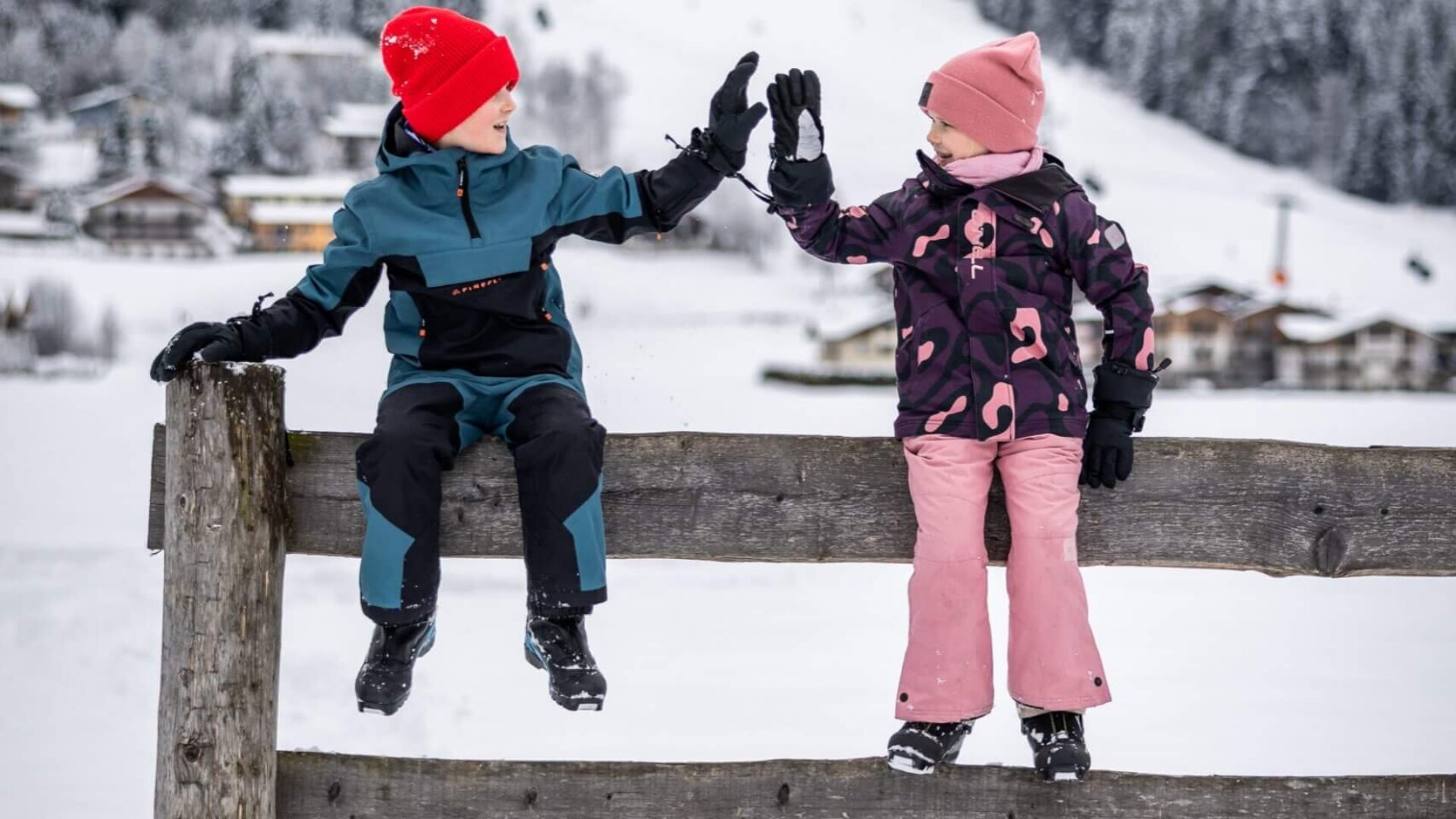 Zwei Kinder sitzen auf Holzzaun im Schnee und geben sich ein High-Five