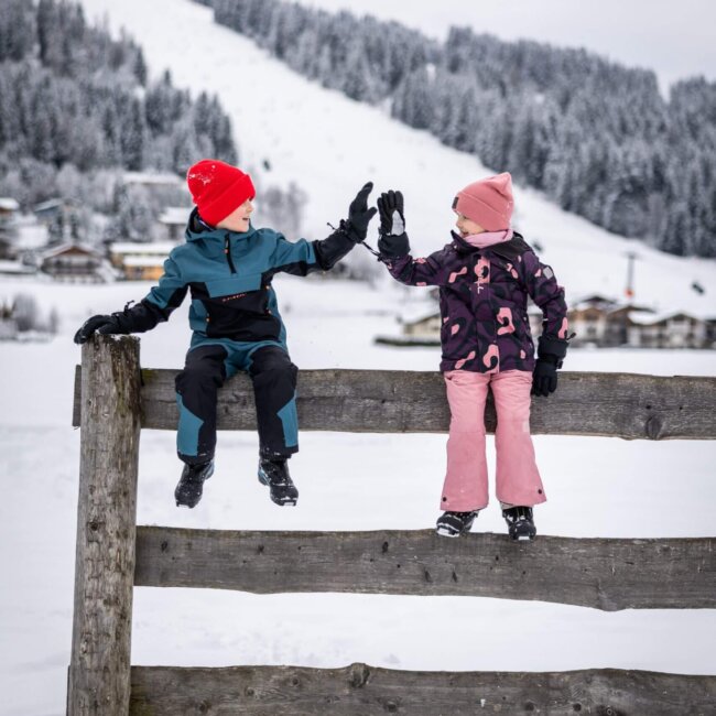 Zwei Kinder sitzen auf Holzzaun im Schnee und geben sich ein High-Five