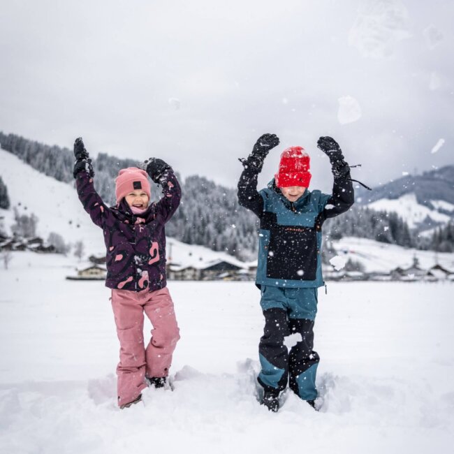 Zwei Kinder spielen lachend im Schnee und werfen Schnee in die Luft