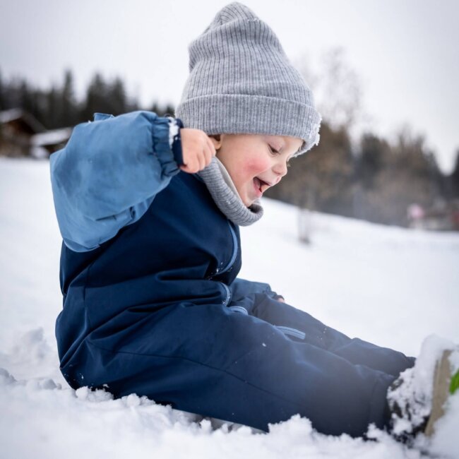 Ein kleines Kind mit Winterkleidung und Mütze sitzt lachend im Schnee.